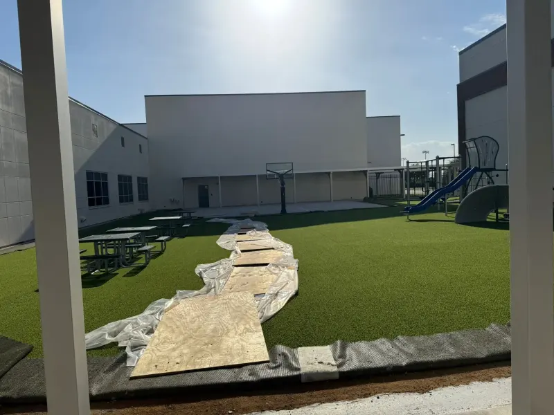 The courtyard mid-build, plywood sheets and sheeting laid across new artificial turf beside the playground and basketball hoop