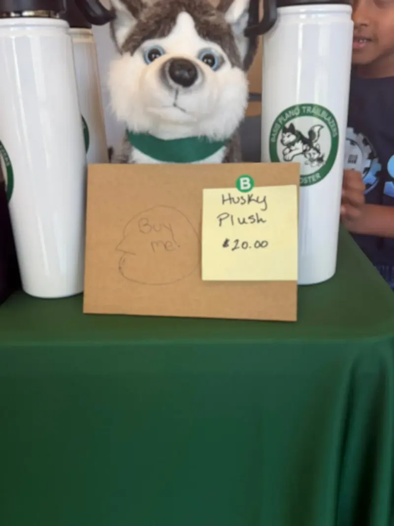A husky plush peering over a hand-lettered “Buy me!” card on the stall, water bottles either side