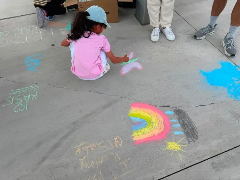 A child in a pink T-shirt and cap kneeling on the walkway to finish a chalk butterfly, a rainbow and sun drawn beside it