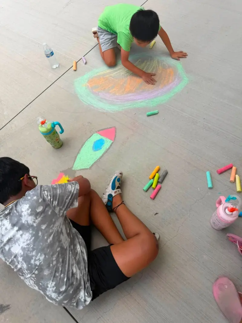 Two children drawing a chalk rocket and a large oval on the path, fistfuls of chalk and water bottles around them