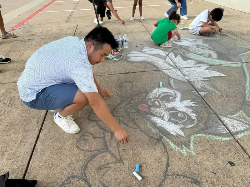 A parent kneeling to outline a large chalk husky while children work on drawings further down the path
