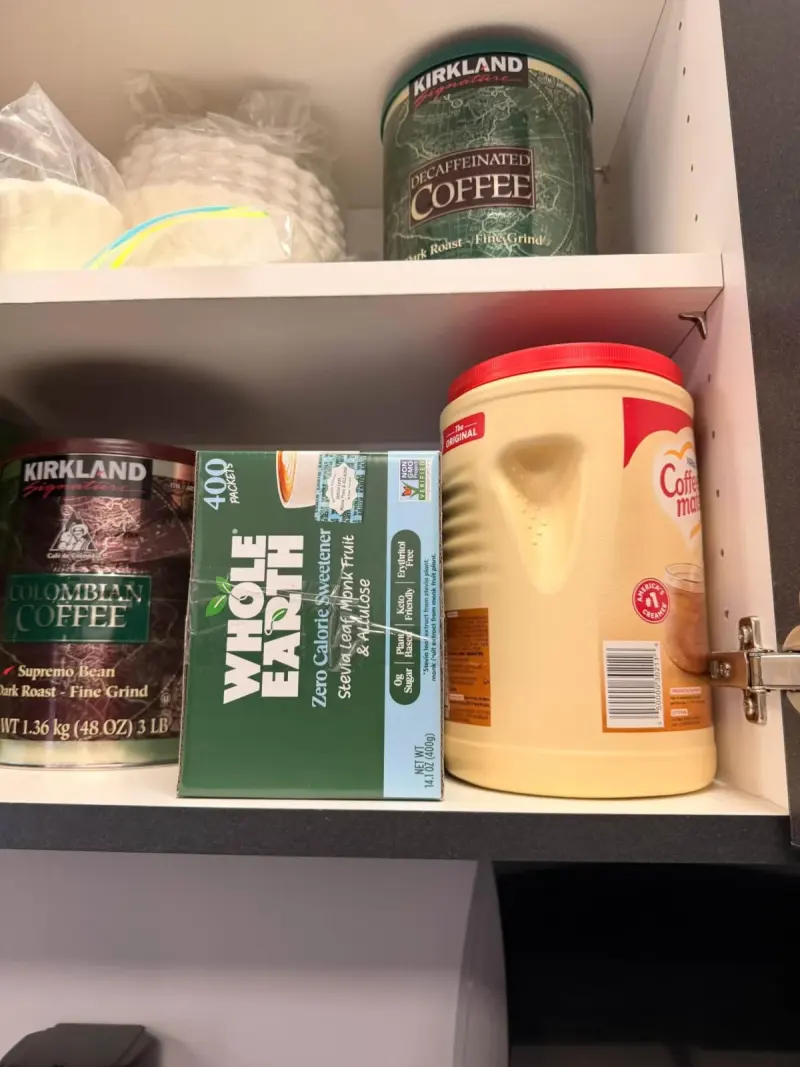 A restocked lounge cupboard: tins of Colombian and decaffeinated coffee, a box of sweetener and a tub of creamer, filters on the shelf above