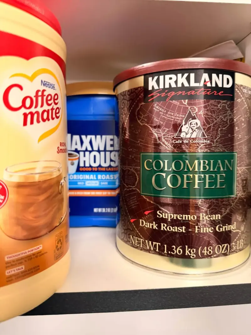 Creamer, instant coffee and a tin of ground Colombian coffee lined up along the cupboard shelf