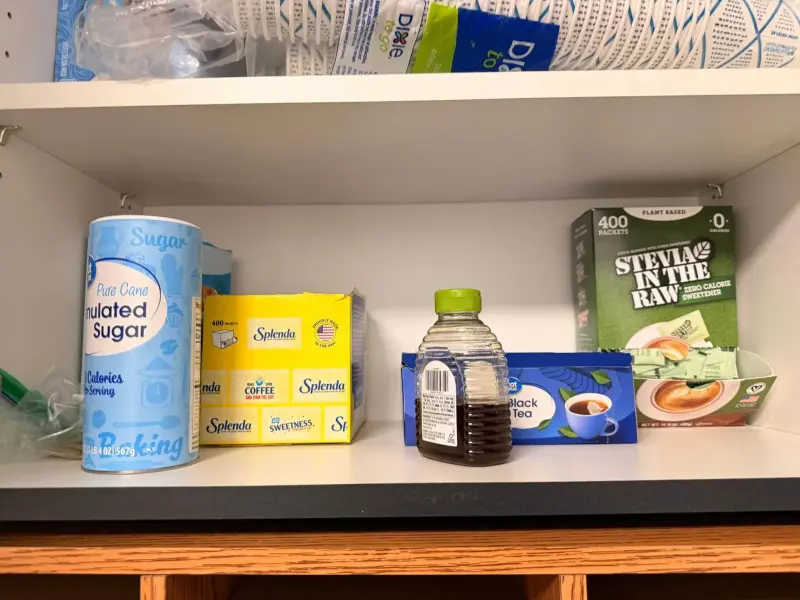 The sweetener shelf restocked with sugar, sachets, honey and a box of black tea, cups stacked above