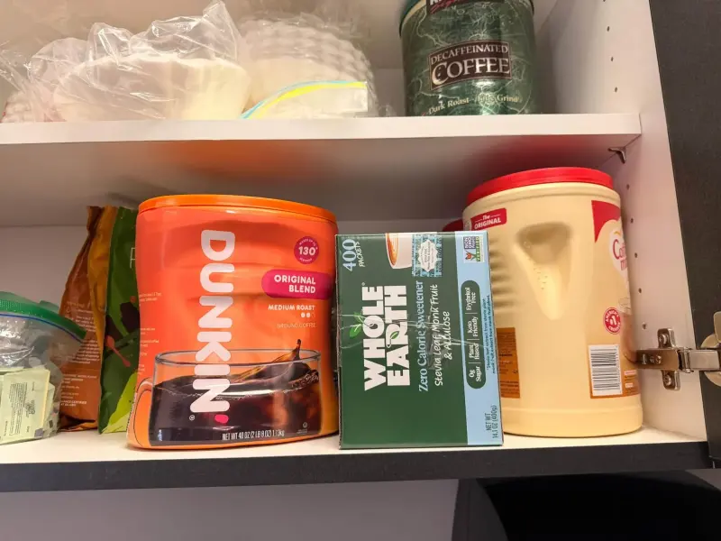 A tub of ground coffee, a box of sweetener and creamer filling the lower shelf, filters and decaf above