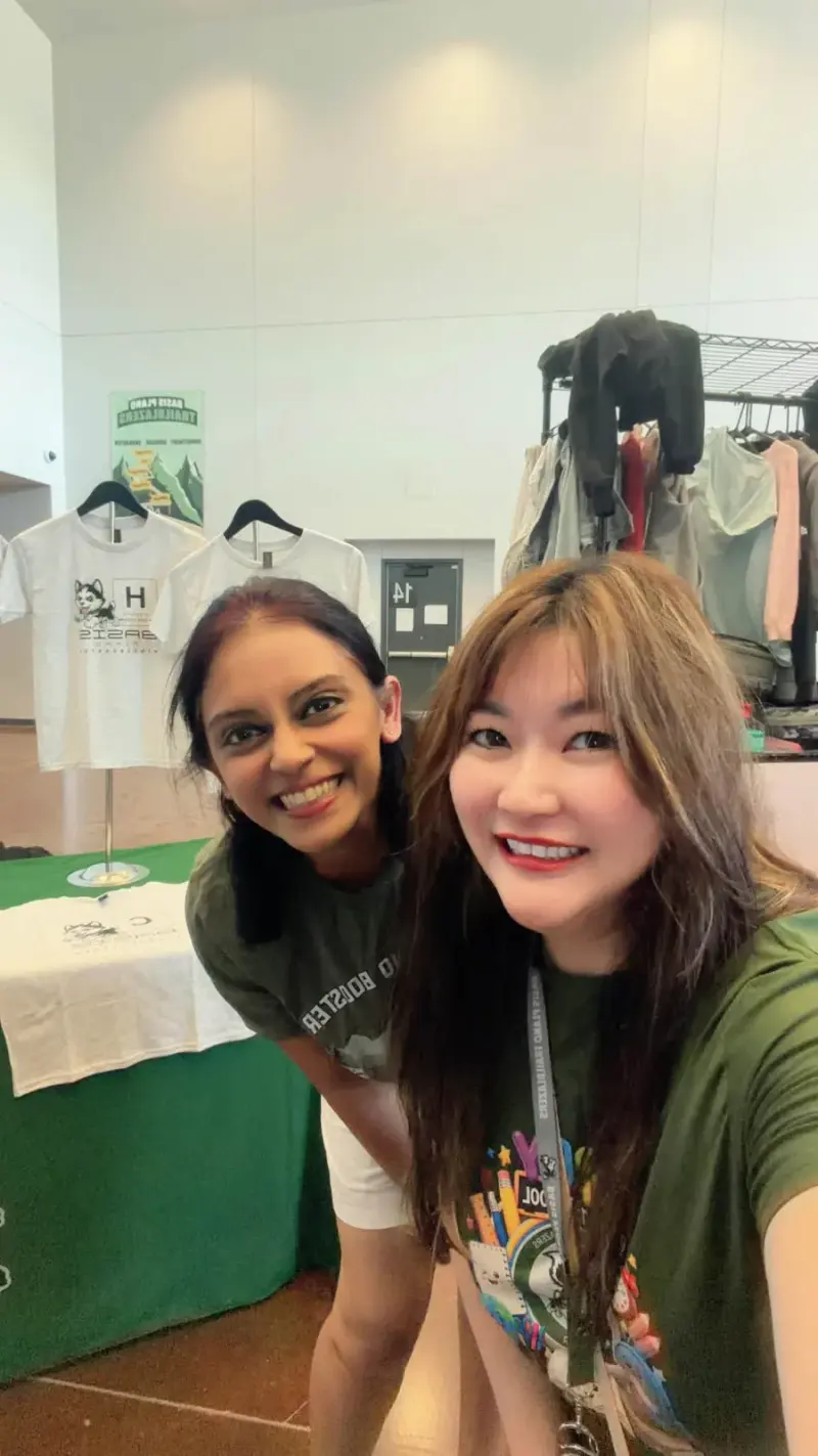 A selfie of two volunteers in Booster shirts in front of the spirit wear table, element shirts on stands behind them