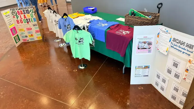 The merchandise table from above, coloured element shirts laid out beside a board of QR codes for the grade-level chat groups