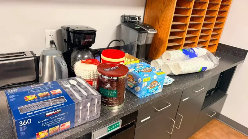 A teacher lounge counter with the coffee maker filled and tubs of coffee, cocoa and cups set out beside it