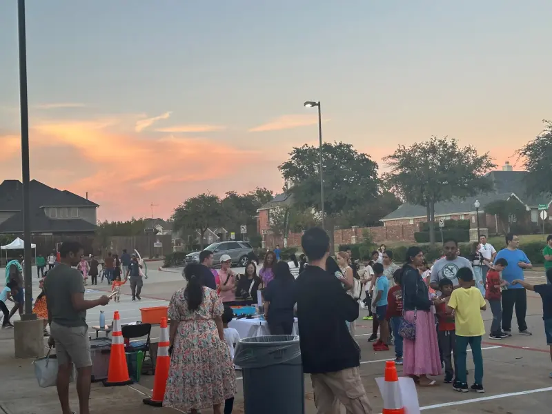 Families crossing the car park between game stalls at dusk, an orange sunset behind the rooftops
