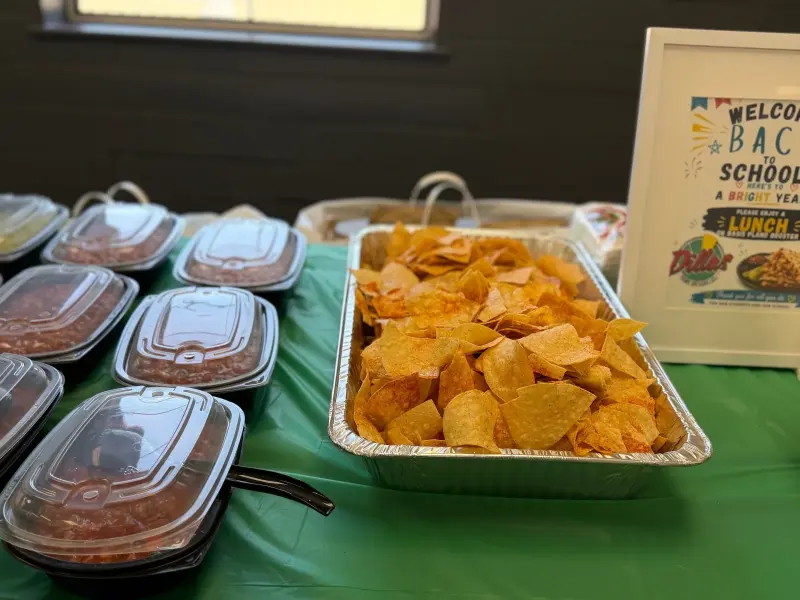 Lidded tubs of salsa and guacamole lined up beside a foil tray of tortilla chips on the green cloth