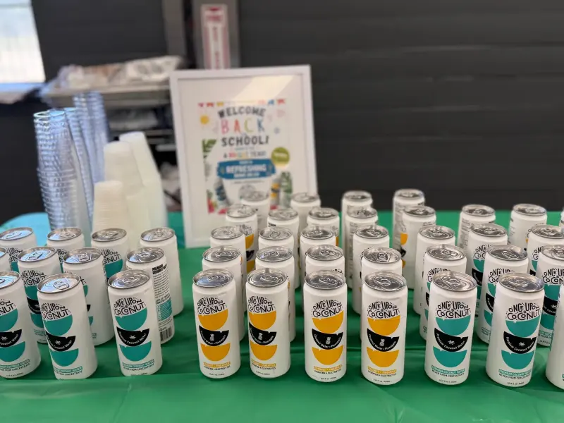 Coconut water cans set out in rows in front of the framed welcome-back sign, cups stacked beside them