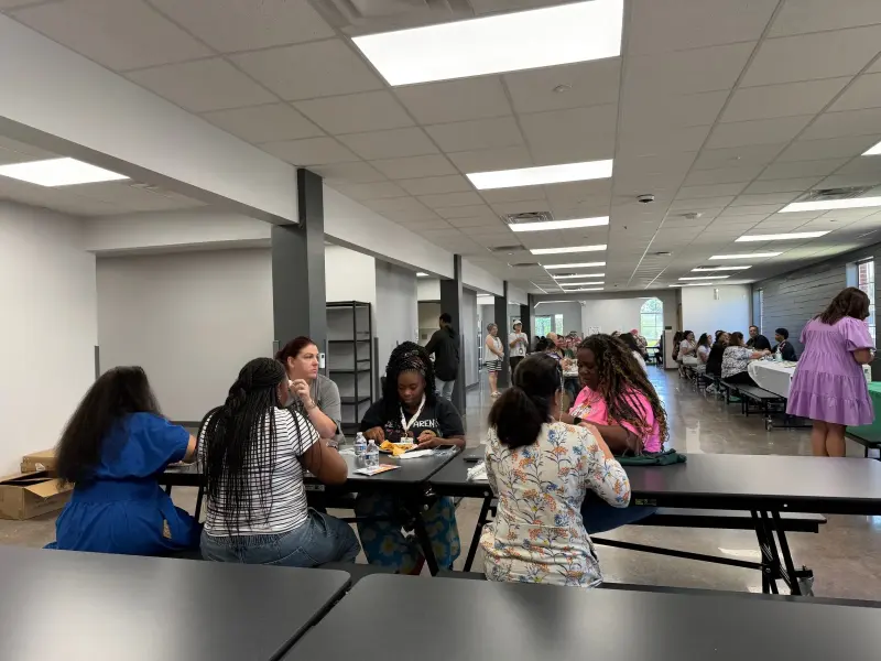 Staff eating and talking around one of the long cafeteria tables, more tables filled behind them