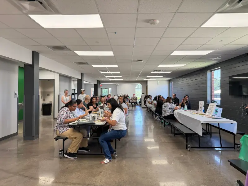 The cafeteria filling up at lunch, staff eating at the long tables with the gift table ready along the far wall