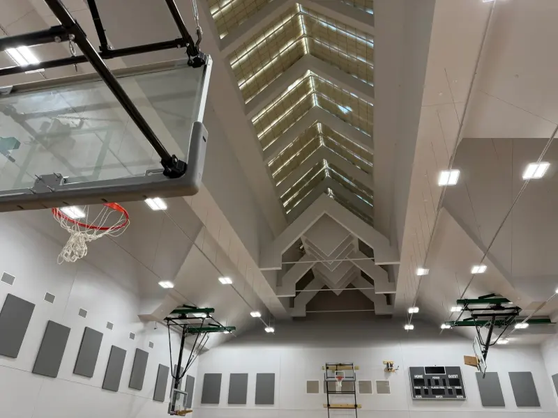 Looking up in the big gym at the peaked clerestory ceiling, a glass backboard in the foreground and the scoreboard on the far wall