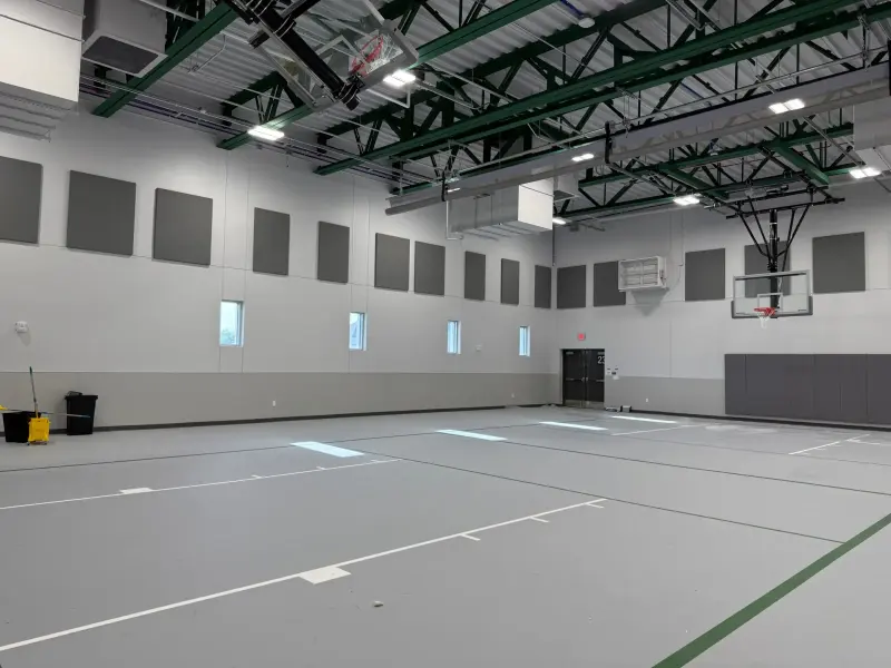 The empty small gym before opening day, grey sport floor freshly marked and backboards raised to the green roof trusses