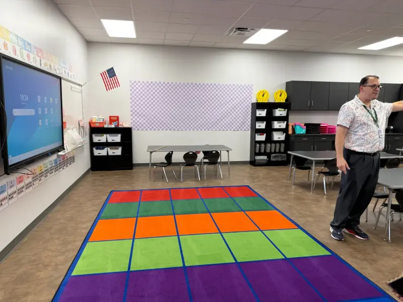 A staff member showing an empty elementary classroom, a block-coloured carpet on the floor and an interactive whiteboard on the wall