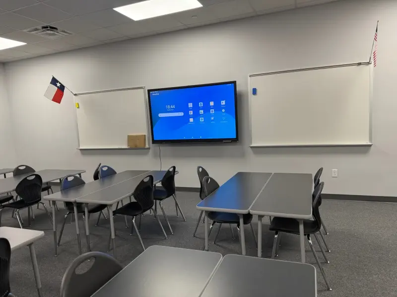 An empty upper-grade classroom with tables and chairs grouped in fours, a display screen between two whiteboards