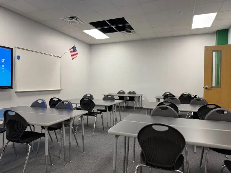 An empty classroom with tables pushed into pairs, a flag on the wall and a display screen beside the whiteboard