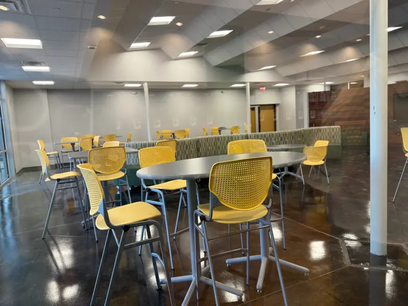 The student commons from the window side, yellow stools and oval tables reflected in the polished floor