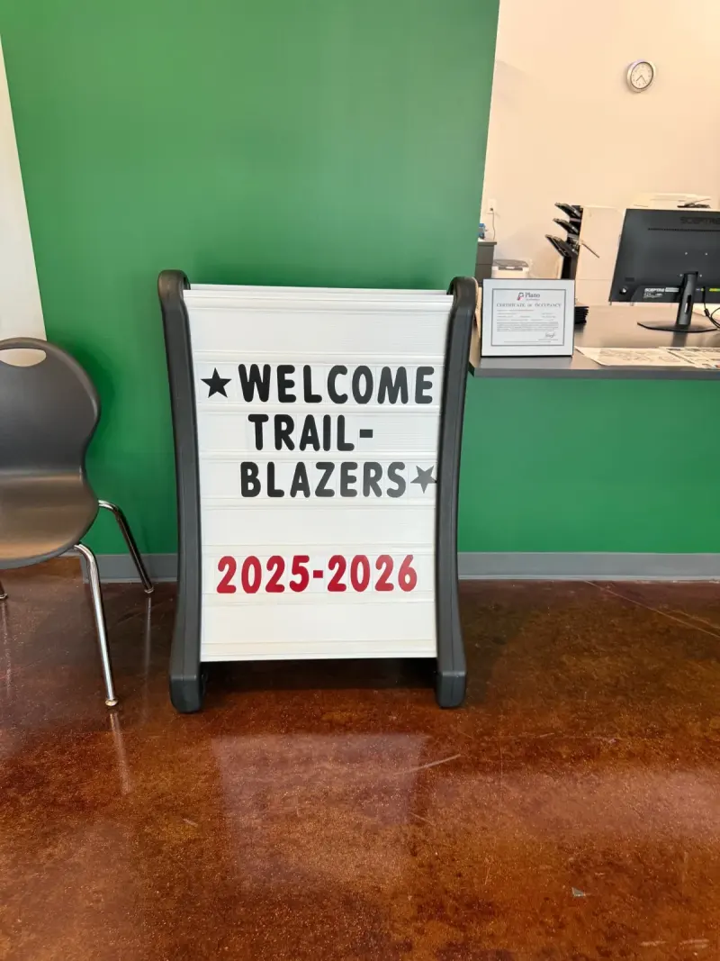 A lobby sign board spelling out “Welcome Trailblazers 2025-2026” in black and red letters against the green front-office wall