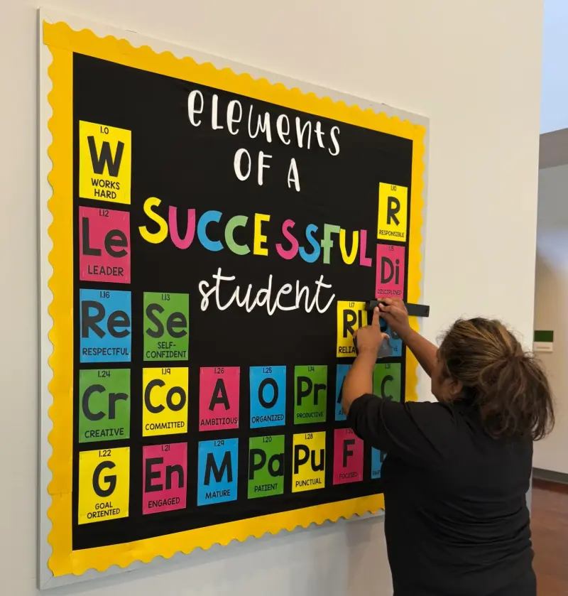 A volunteer pinning the last letter tiles onto an “Elements of a Successful Student” bulletin board