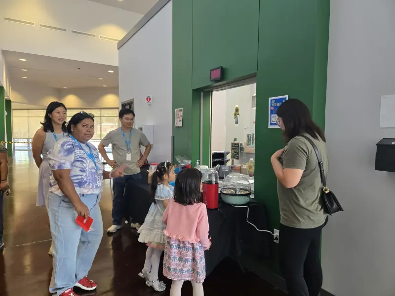 Booster volunteers on visitor lanyards talking beside the concession table while two children wait for a fresh batch of popcorn