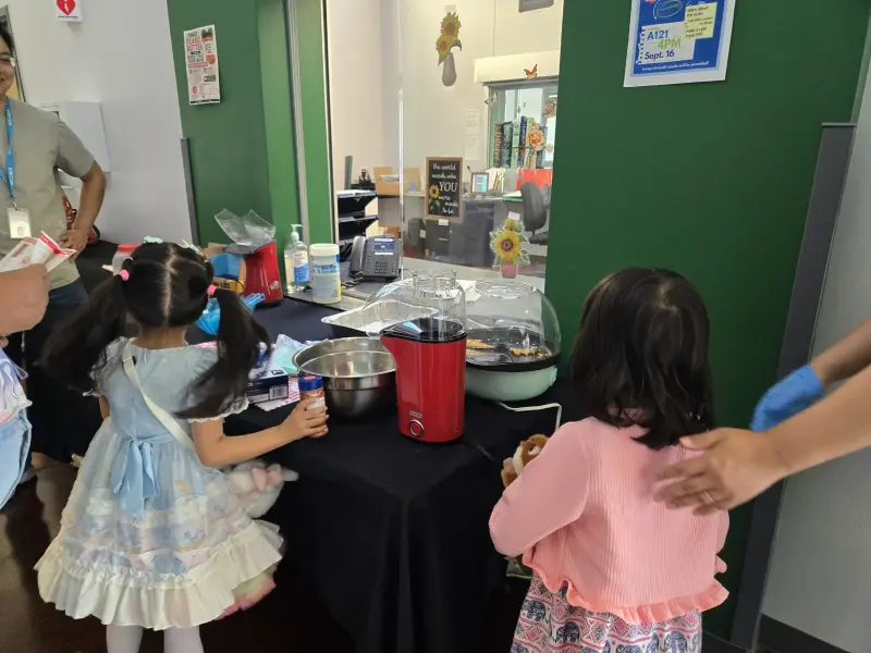Two children at the concession table watching the red popcorn maker, mixing bowls and a jar of kernels set out beside it
