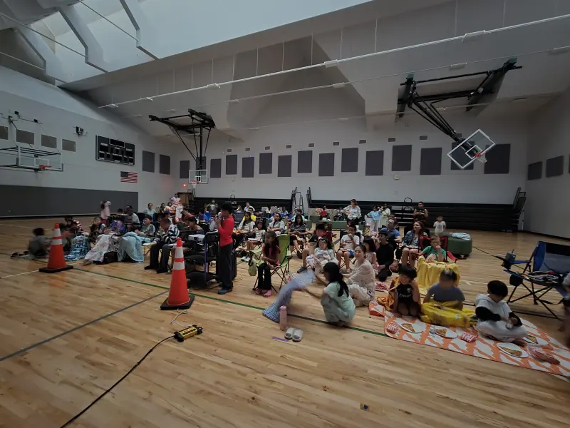 Families spread across the gym floor on blankets and folding chairs with plates of pizza beside them, watching the screen