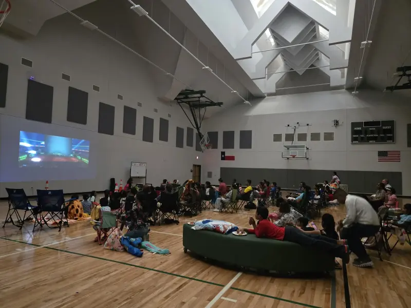 A wide view down the gym as the movie plays, folding chairs and cushioned benches filled with families