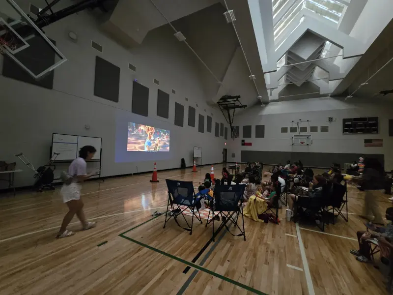 A volunteer crossing the gym floor while the film plays, families gathered in chairs on the far side