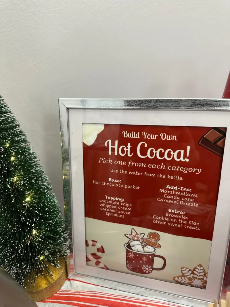 A framed “Build Your Own Hot Cocoa!” card listing base, add-ins, toppings and extras, a lit mini tree beside it