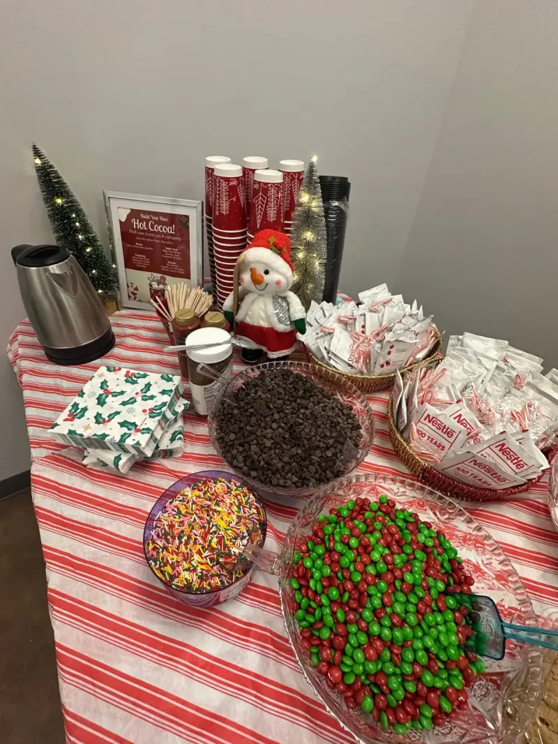 A hot cocoa bar on a candy-striped cloth: kettle, red cups, a bowl of chocolate chips, sprinkles and red and green sweets
