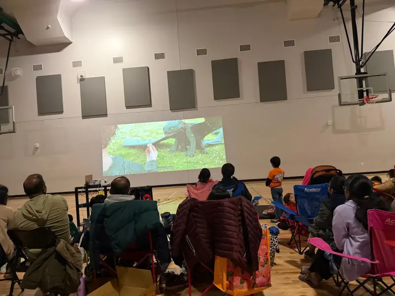 Families in camping chairs and blankets watching an animated dragon on the projected screen in the darkened gym