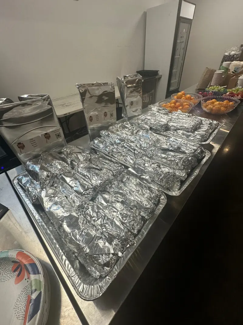Foil-wrapped breakfast burritos filling two large trays on a stainless counter, with bowls of clementines beside them
