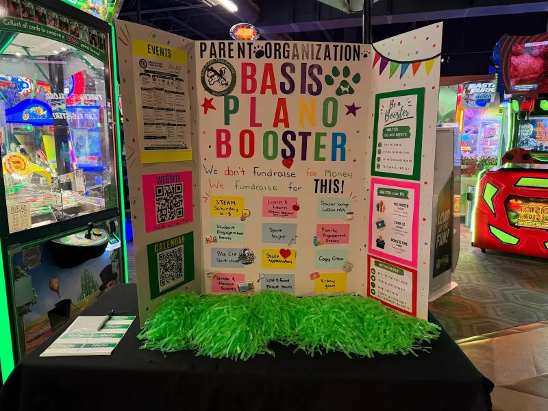 The Booster’s trifold display board at the arcade, headed “We don’t fundraise for money, we fundraise for THIS!” with QR codes and event cards, green pom-poms along the front of the table