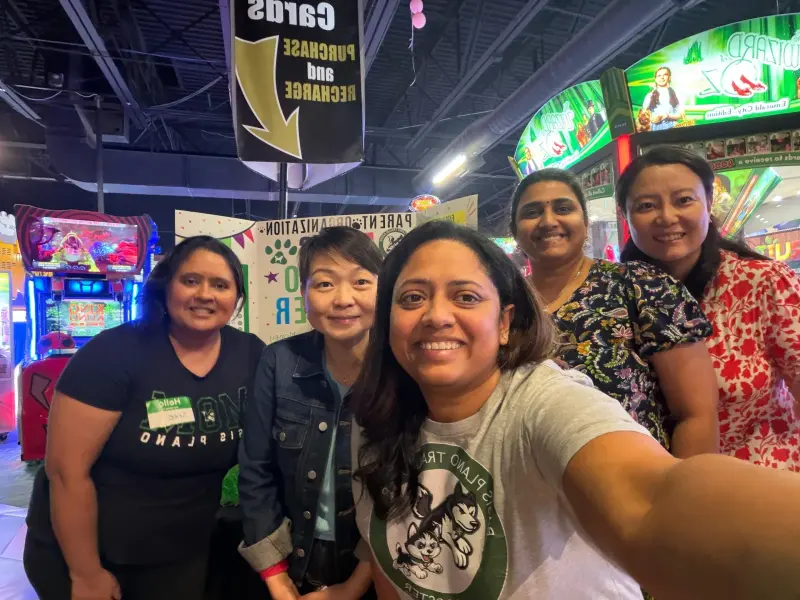 A selfie of five volunteers crowded in front of the Booster board, arcade machines lit up behind them