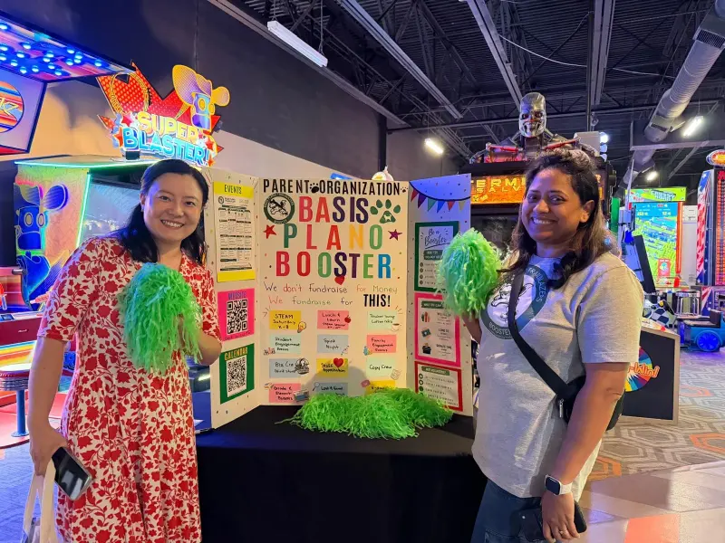 Two volunteers holding green pom-poms either side of the Booster display board, arcade cabinets on both sides
