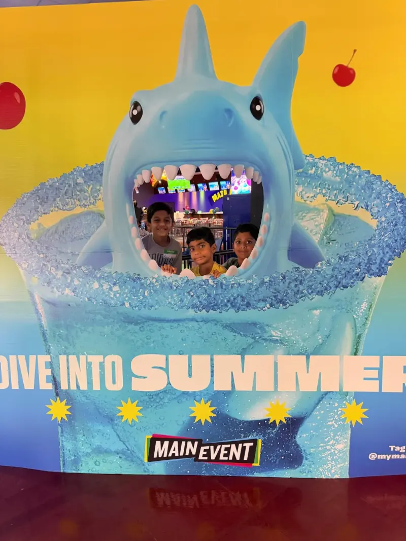 Three children grinning out through the mouth of a giant shark cut-out on a “Dive into Summer” photo board